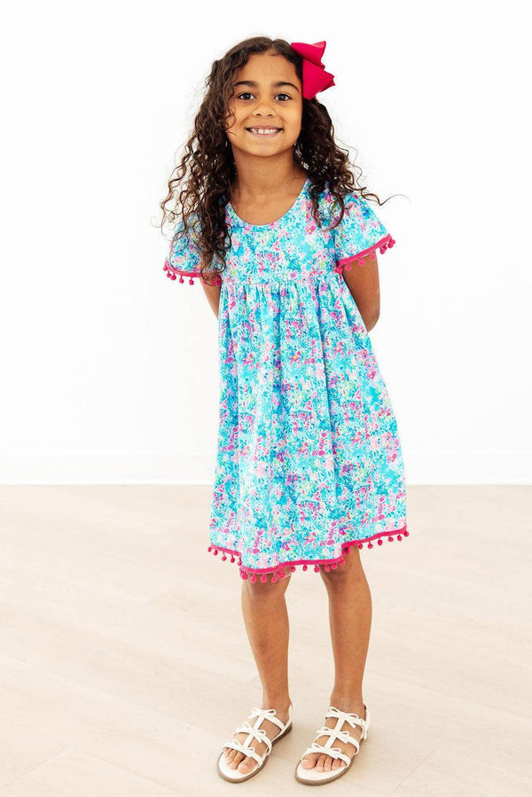 Aqua Garden Pom Pom Dress in blue floral print with pink pom pom trim on sleeves and hem worn by a smiling girl