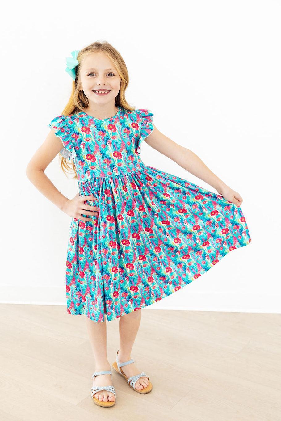 Girl in Lovely Llamas Flutter Sleeve Twirl Dress, teal floral print with flutter sleeves and a twirling skirt.