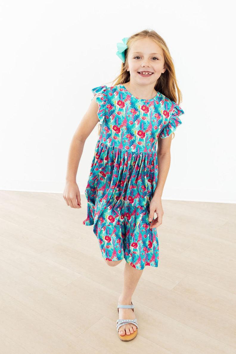 Girl wearing Lovely Llamas Flutter Sleeve Twirl Dress in blue floral print with flutter sleeves and turquoise bow.