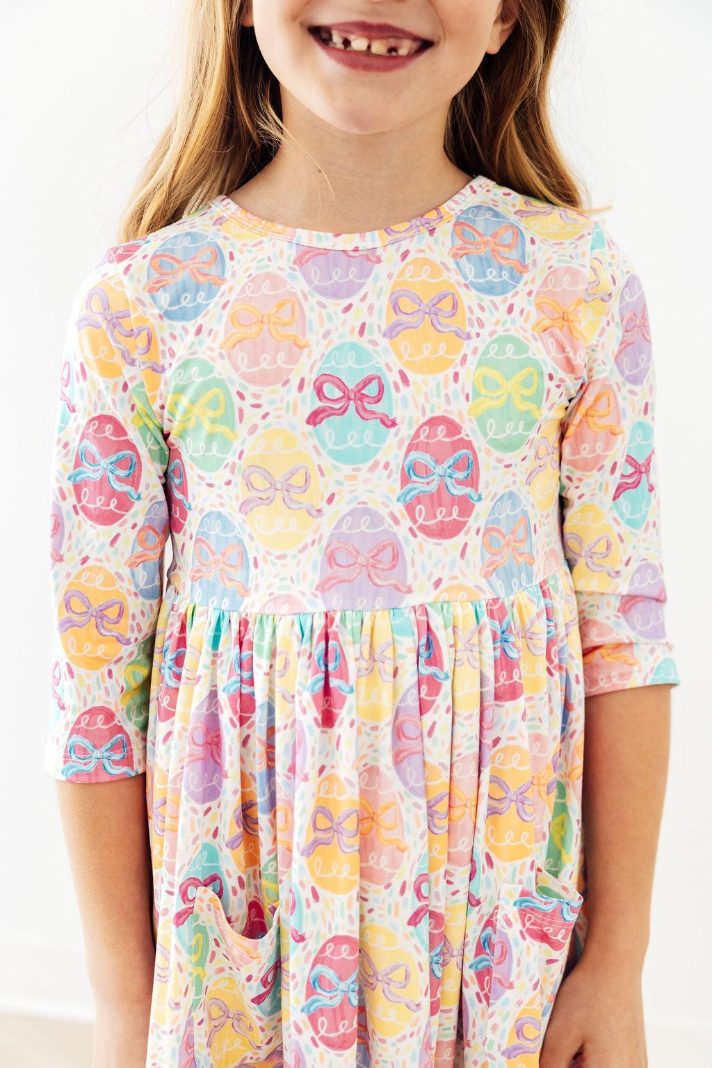 Smiling toddler in pastel egg and bow print twirl dress with 3/4 sleeves.
