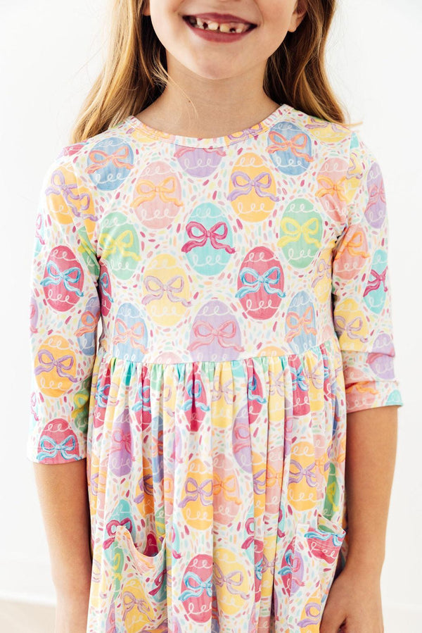 Smiling toddler in pastel egg and bow print twirl dress with 3/4 sleeves.