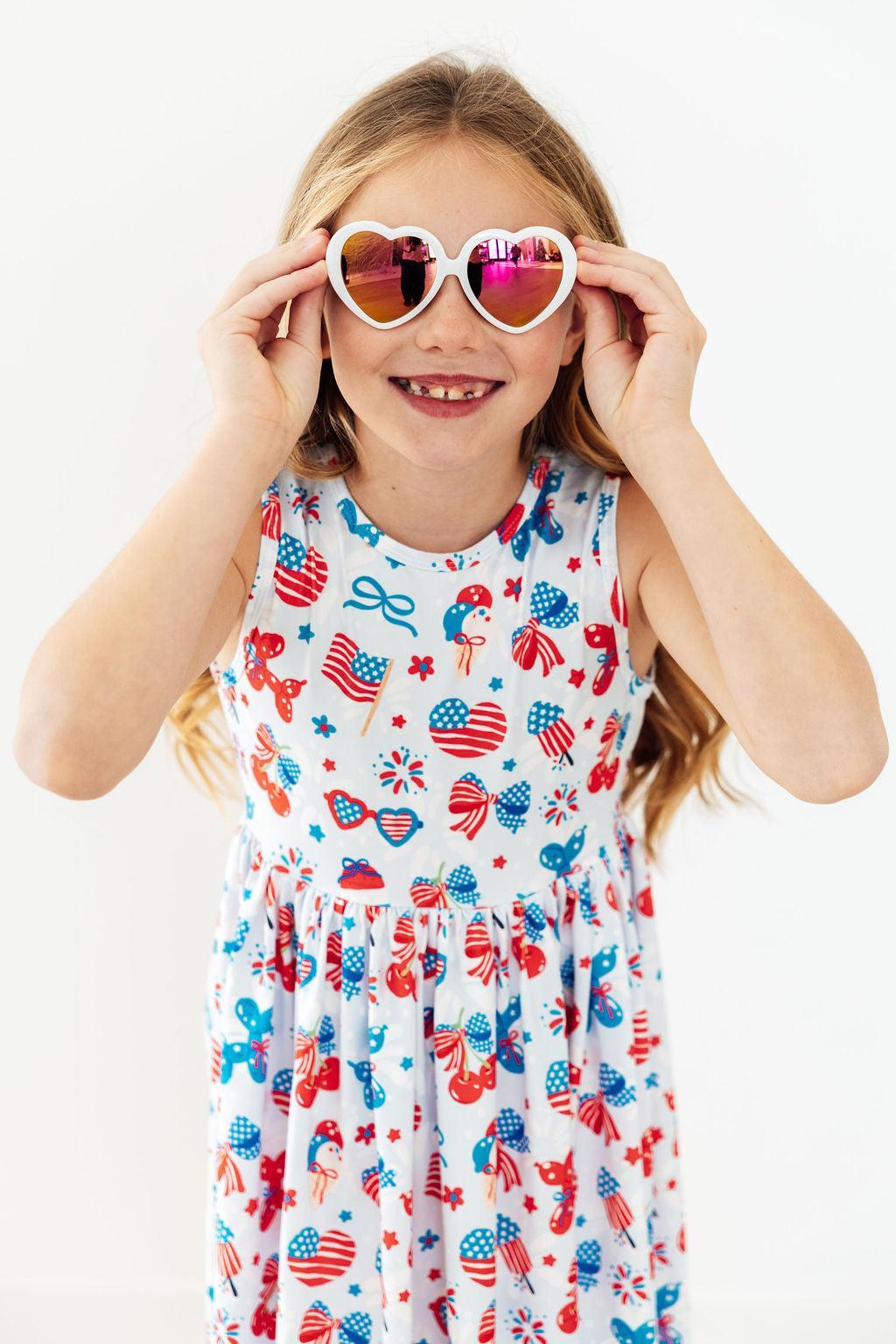 Girl wearing Little Miss Liberty Tank Twirl Dress in red, white, and blue with heart sunglasses.