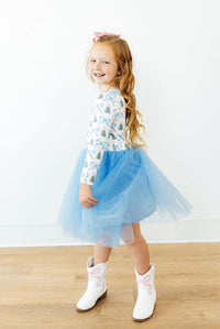 Girl in white long sleeve Christmas print top with blue tutu skirt and white cowboy boots featuring pink bows.