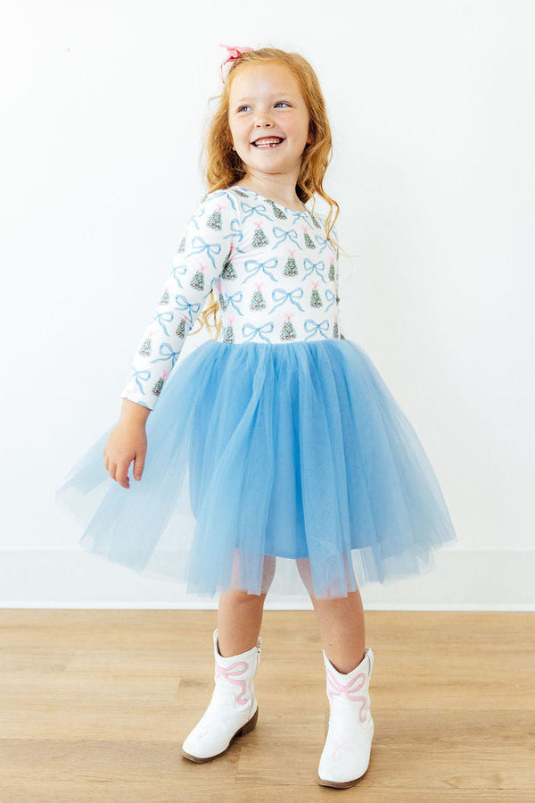 Girl wearing Christmas long sleeve tutu dress with blue skirt and tree print top, paired with white cowboy boots.