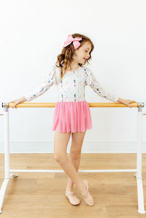 Young ballerina at a barre wearing a long sleeve holiday leotard with a pink tulle skirt and matching bow. Perfect for dance class.