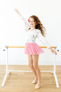 Young ballerina posing at a barre in a long sleeve Christmas print leotard with a pink tulle skirt.