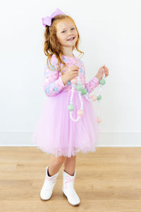 Girl wearing pink long sleeve tutu dress with light-up print and white cowboy boots, holding a pastel bead necklace.