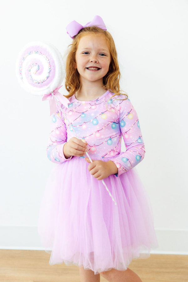 Girl in pink long sleeve tutu dress with string lights print holding a large lollipop prop and wearing a purple bow.