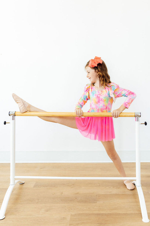 Child stretching at ballet barre wearing colorful tie-dye long sleeve twirl leotard with pink skirt and orange bow.