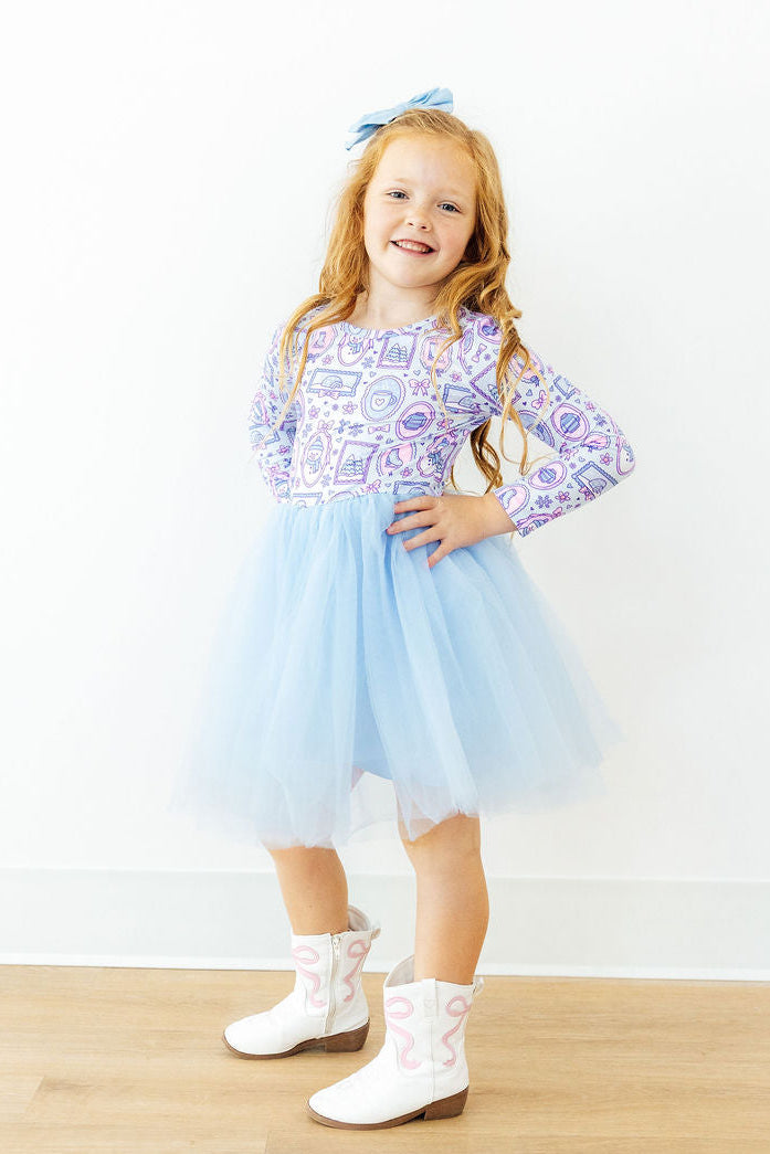 Girl in long sleeve tutu dress with winter print bodice and light blue tulle skirt, paired with white cowboy boots and blue bow.