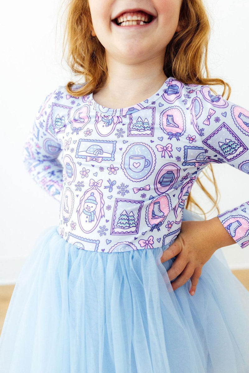 Girl wearing long sleeve tutu dress with winter print featuring ice skates, snowmen, and trees, paired with a light blue tulle skirt.