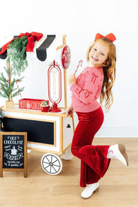 Girl in pink bow print sweater and red velvet flared pants holding a candy cane near a hot cocoa cart.