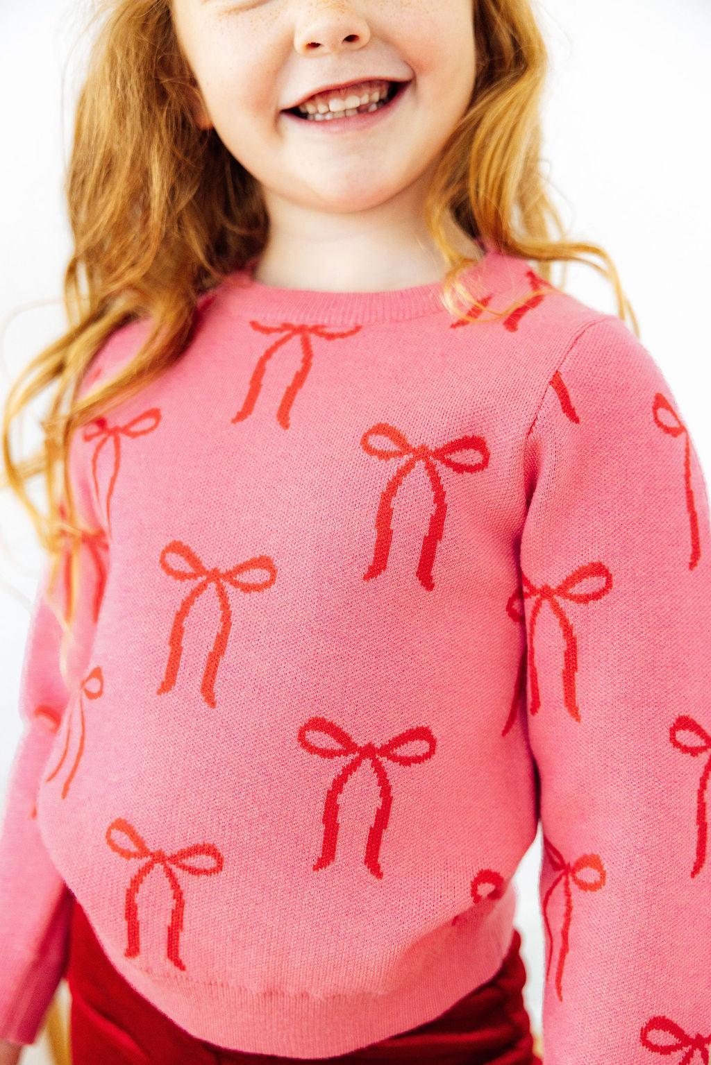 Pink sweater with red bow pattern for girls. Soft, true-to-size knit perfect for layering over dresses or wearing with leggings on chilly days.