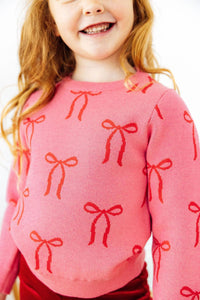 Pink sweater with red bow pattern for girls. Soft, true-to-size knit perfect for layering over dresses or wearing with leggings on chilly days.