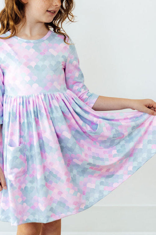 Child holding the full skirt of a pastel mermaid scale print 3/4 sleeve twirl dress with pockets.