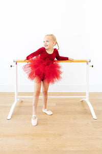 Little girl in a red velvet long sleeve leotard with a fluffy分层 tutu skirt posing at a ballet barre.