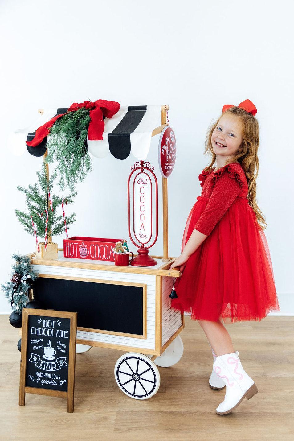 Girl smiling in a red shimmer 3/4 sleeve tutu dress with flutter sleeves and white cowboy boots beside a hot cocoa cart.