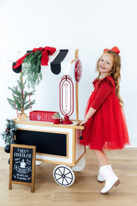 Girl smiling in a red shimmer 3/4 sleeve tutu dress with flutter sleeves and white cowboy boots beside a hot cocoa cart.