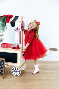 Happy girl in a red shimmer 3/4 sleeve tutu dress with flutter sleeves and white cowboy boots posing by a hot cocoa cart.