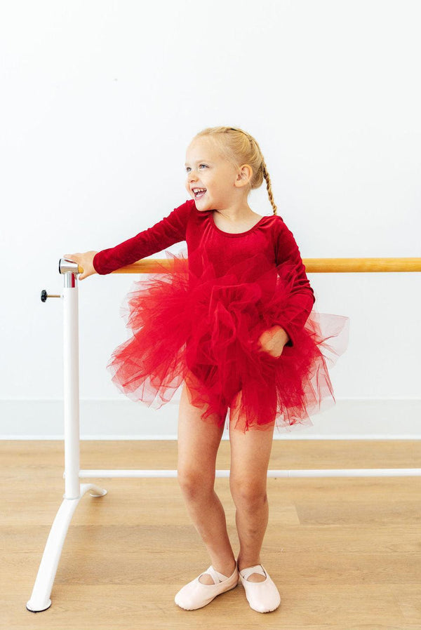 Happy ballerina girl wearing a red velvet long sleeve leotard with a fluffy multi-layer tulle tutu skirt at the dance barre.