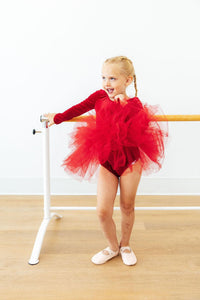 Little girl in a red velvet long sleeve tutu leotard posing at a ballet barre with fluffy tulle skirt and pink shoes.
