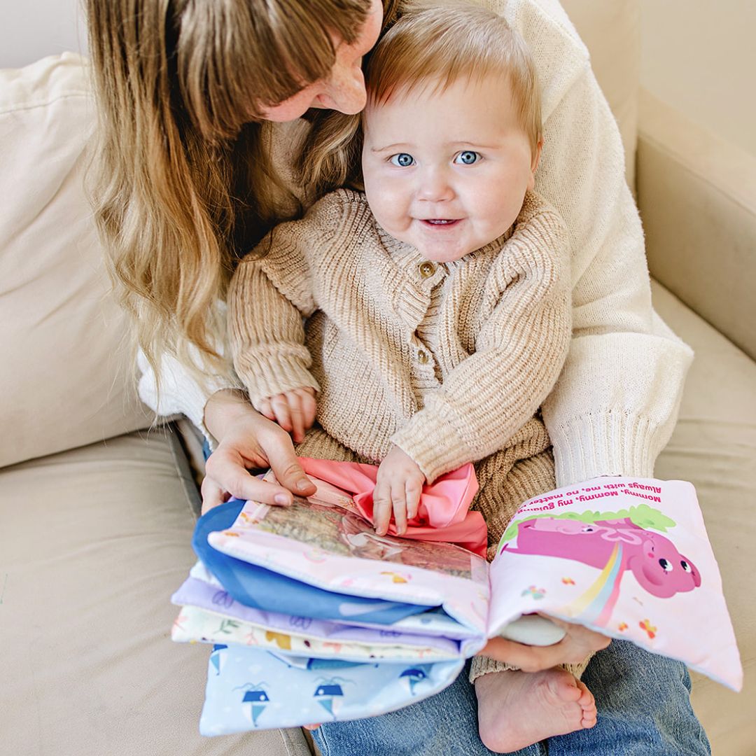 Baby in beige knit sweater and pink pants with adult holding colorful Peek-A-Photo interactive books using 