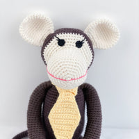 Handmade brown amigurumi monkey with cream ears and a yellow tie, featuring a pink smile for nursery decor or pretend play.