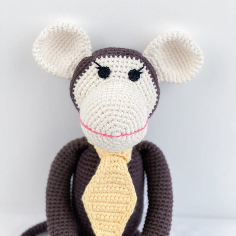 Handmade brown amigurumi monkey with cream ears and a yellow tie, featuring a pink smile for nursery decor or pretend play.