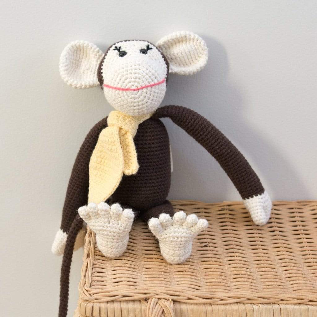 Handmade brown crochet monkey with cream face, yellow scarf, and long tail sitting on a wicker basket.