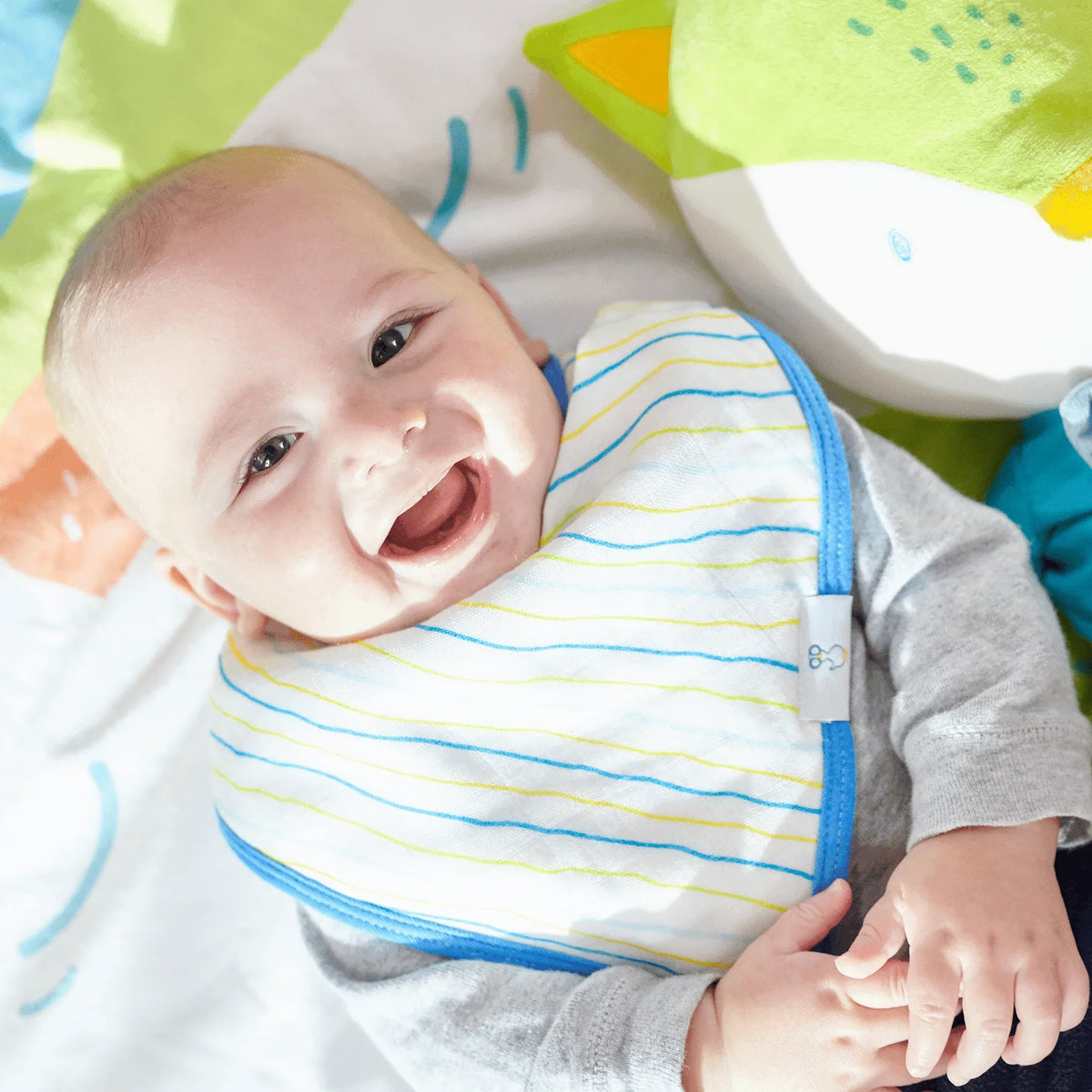 Smiling baby wearing blue trimmed muslin and terry cloth baby bib set lying on colorful blanket