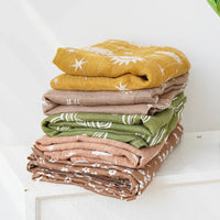 Stack of colorful muslin swaddle blankets in mustard, beige, green, and brown with white patterns for babies
