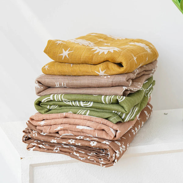 Stack of colorful muslin swaddle blankets in mustard, beige, green, and brown with white patterns for babies