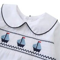 Close-up of white nautical smocked romper for baby boys with blue sailboat embroidery and black trim collar