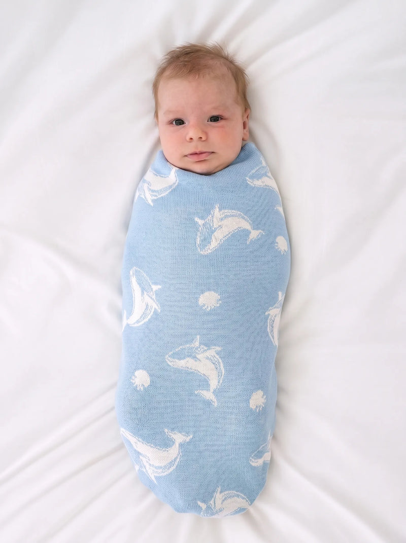 Newborn swaddled in light blue whale patterned knit blanket on white textured sheet showing soft ribbed texture