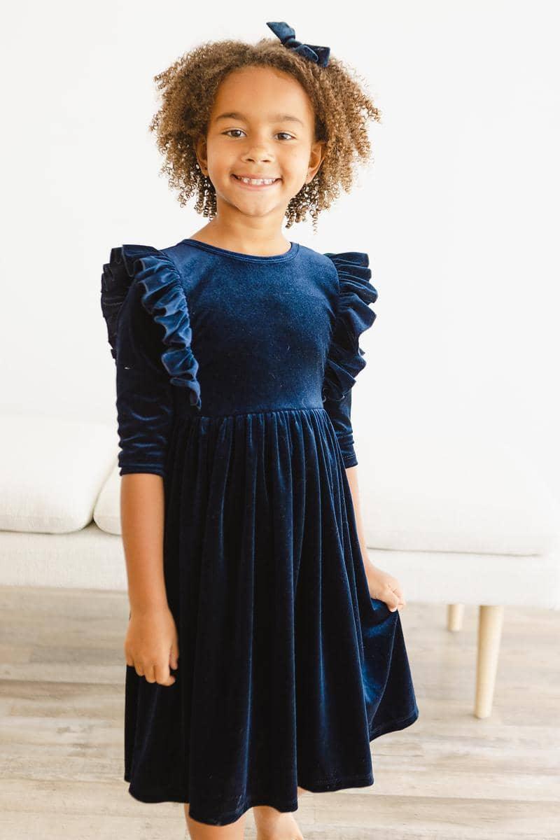 girl wearing navy velvet ruffle dress for girls standing and smiling indoors