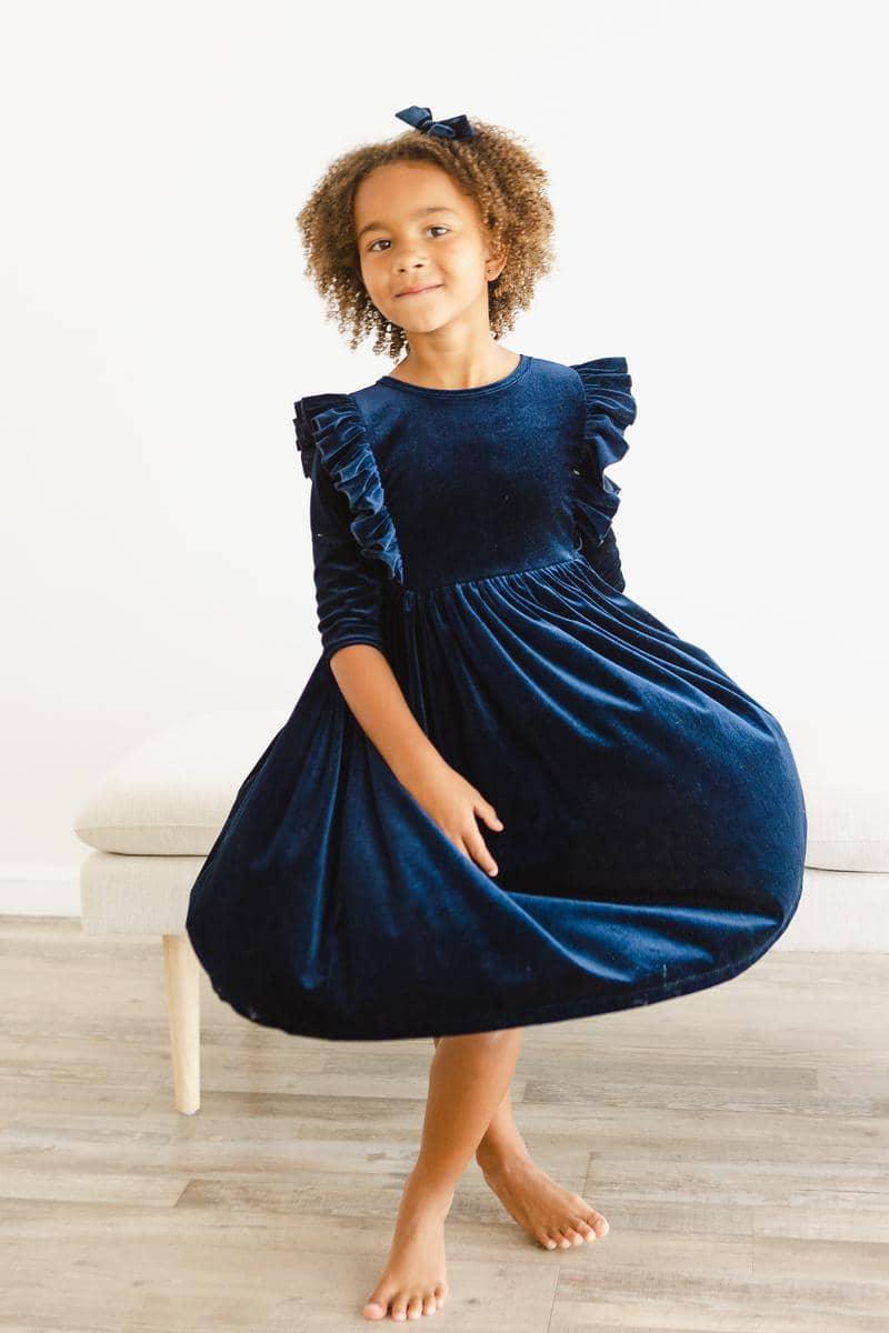 girl wearing navy velvet ruffle dress for girls twirling indoors barefoot with matching hair bow