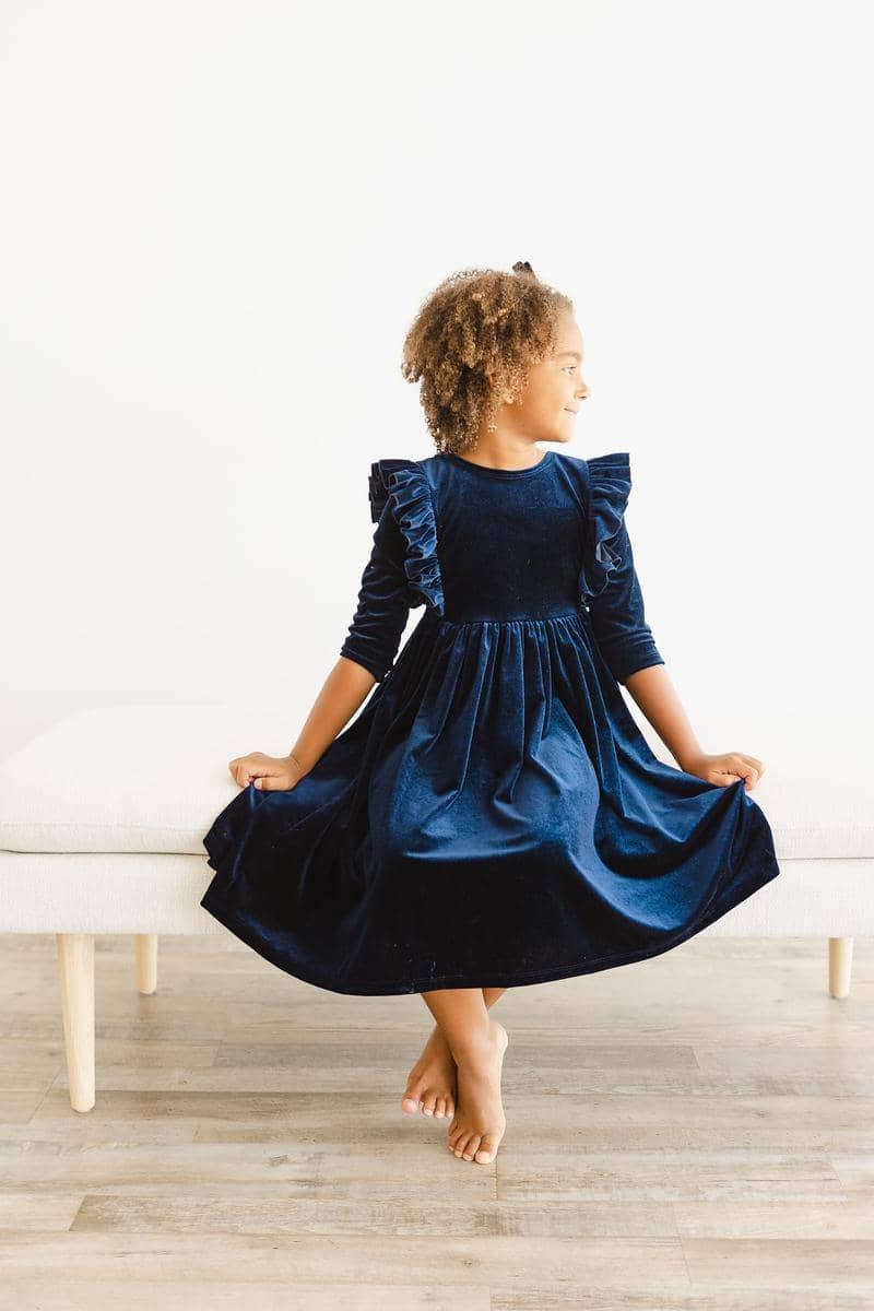 Girl wearing navy velvet ruffle dress for girls sitting on bench holding dress hem