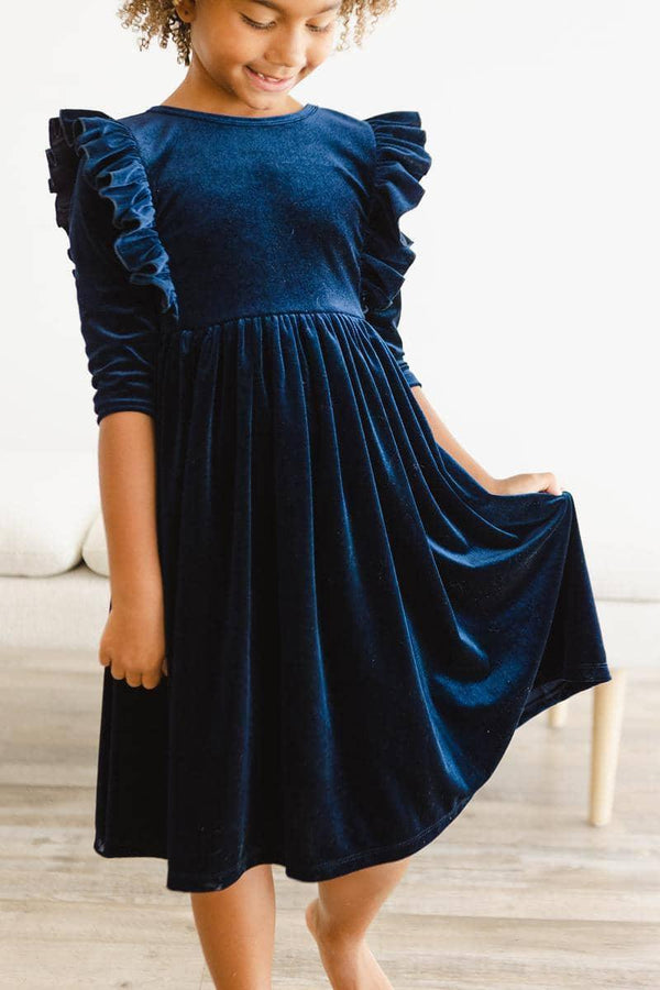 young girl wearing navy velvet ruffle dress for girls holding the skirt while smiling indoors