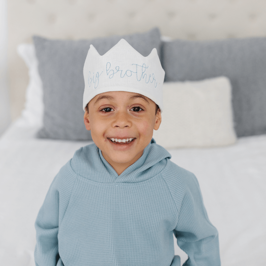 Child wearing a white crown with text, sitting on a bed with gray pillows.