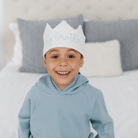 Child wearing a white crown with text, sitting on a bed with gray pillows.