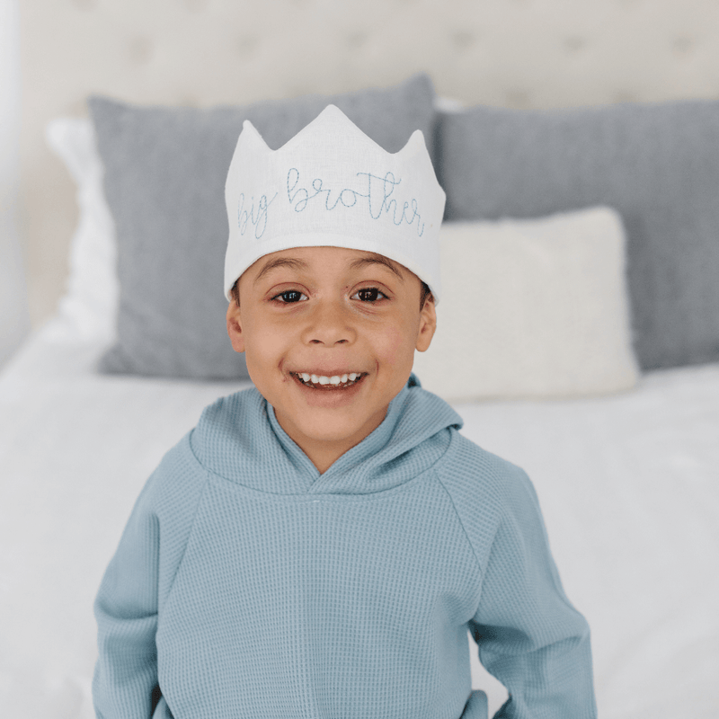 Child wearing a white crown with text, sitting on a bed with gray pillows.