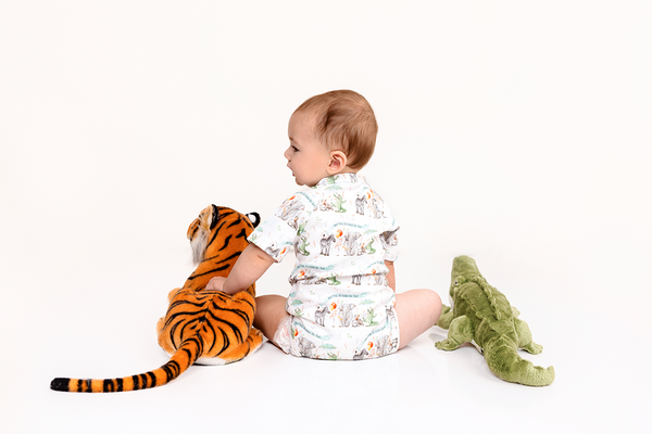 Baby wearing an organic cotton onesie with animal print sits with tiger and crocodile plush toys.