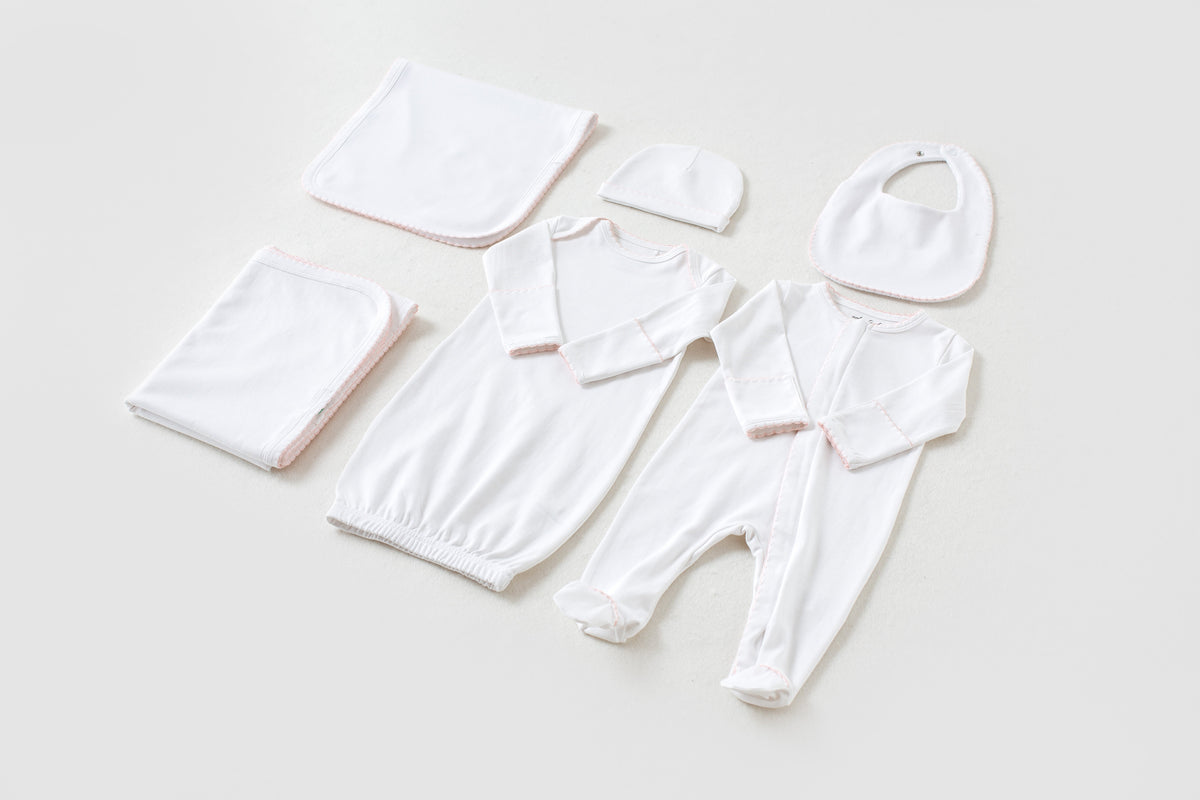 White newborn take me home set with pink trim, including gown, footed pajama, hat, bib, and blankets.