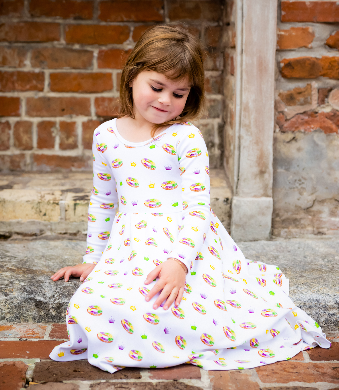 Young girl sitting on a stone step wearing a white dress with colorful patterns against a brick wall.