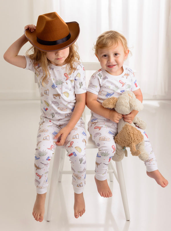 Two kids wearing Texas Kids Organic Cotton Sleepwear with desert and travel prints, cuddling a teddy bear.