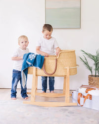 Two boys stand by a wooden bassinet draped with a navy blue baby blanket featuring white sea turtle designs.