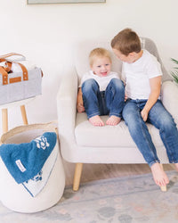 Nursery toy storage basket filled with blue towel next to cream armchair where two boys are sitting