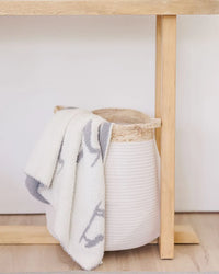 Creamy white baby blanket with light charcoal heron print draped over a woven basket under a wooden table.