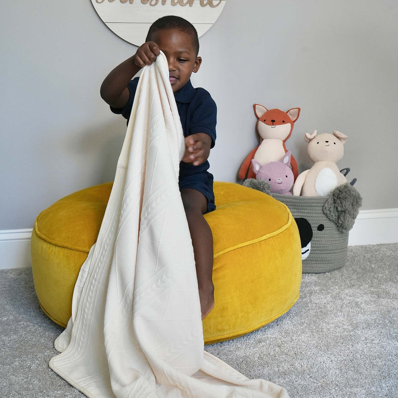 A happy child playing with the Oatmeal Knit Blanket from Goosewaddle®, on a soft yellow pouf, surrounded by plush toys in a cozy nursery setting.