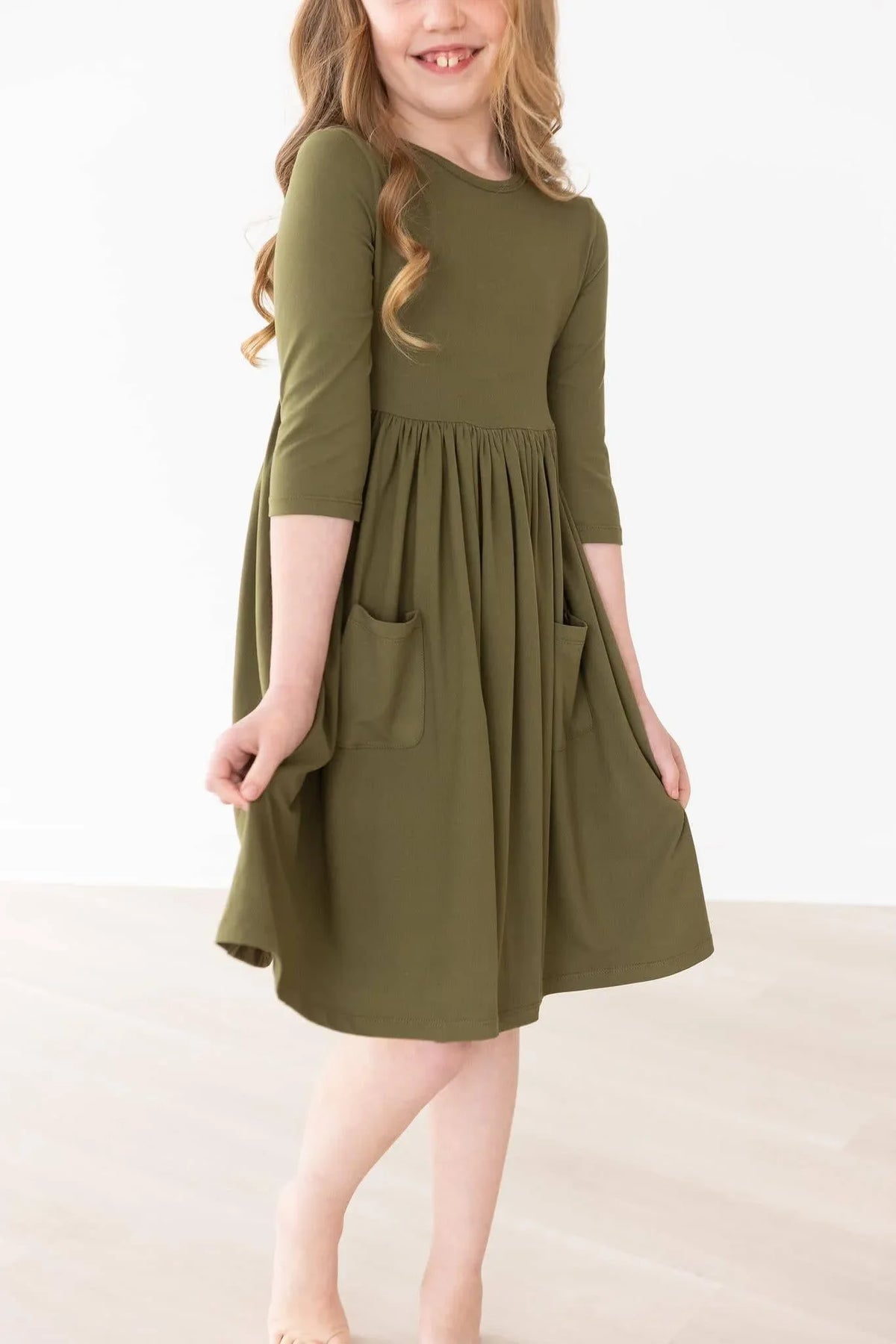 Girl wearing olive twirl dress for girls with 3/4 sleeves and pockets spinning indoors